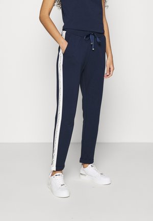 Kvinde iført marineblå joggingbukser med hvide sidestriber og hvide sneakers, stående med den ene hånd i lommen mod en ensfarvet baggrund.