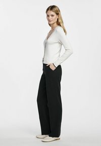 Biały długi kardigan z długim rękawem, w kształcie litery V i z zamkiem z przodu, zestawiony z czarnymi spodniami o szerokich nogawkach i jasnymi butami. Widok z przodu.