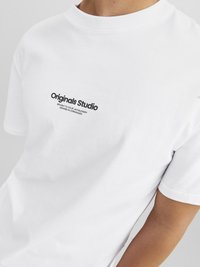 Balta kokvilnas T-kreklu ar apaļu kaklu un īsām piedurknēm. Uz tā ir melns uzraksts "Originals Studio" ar zīmola detaļām zem tā.
