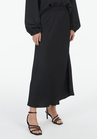 Schwarzer Maxirock aus glattem Stoff mit elastischem Taillenbund, seitlichem Schlitz und kombiniert mit schwarzen, gebundenen High-Heels.