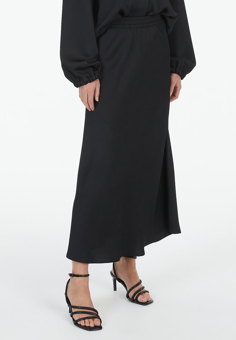 Schwarzer Maxirock aus glattem Stoff mit elastischem Taillenbund, seitlichem Schlitz und kombiniert mit schwarzen, gebundenen High-Heels.