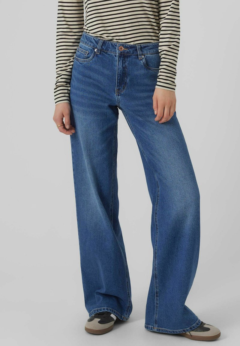 Blaue Denim-Jeans mit weitem Bein, hoher Taille, fünf Taschen und sichtbaren Nähten. Kombiniert mit einem schwarz-weißen gestreiften Langarmoberteil.