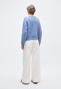 Μπλε πλεκτό πουλόβερ με υφασμάτινες λεπτομέρειες· μακριά μανίκια, στρογγυλή λαιμόκοψη. Συνδυασμένο με φαρδιά κρεμ παντελόνια και μαύρα παπούτσια.