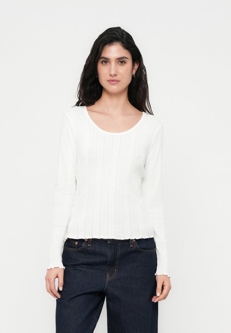 Λευκό μπλουζάκι με μακριά μανίκια, στρογγυλή λαιμόκοψη, υφασμάτινη ριμπ υφή και σκάλιστο τελείωμα, συνδυασμένο με σκούρα, χαλαρά τζιν.