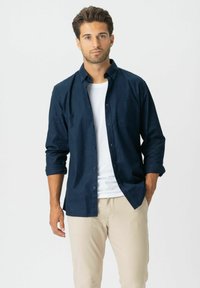 Man met kort donker haar, gekleed in een marineblauwe overhemd met knopen over een wit T-shirt en beige broeken, staand tegen een witte achtergrond.