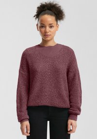 Pull à col en V et à manches longues en fil bordeaux texturé. Coupe ample avec des manches larges, superposé sur un haut bordeaux. Un pantalon noir complète la tenue.