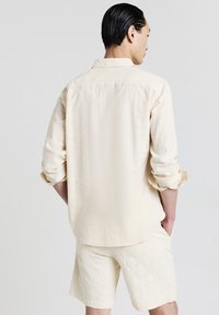 Uomo di spalle con una camicia a maniche lunghe color crema con texture e maniche arrotolate, abbinata a pantaloni corti, con le mani nelle tasche.