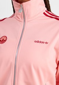 Giacca zip-up Adidas rosa con strisce rosse sulle spalle e loghi sul petto, indossata da una persona con capelli intrecciati.