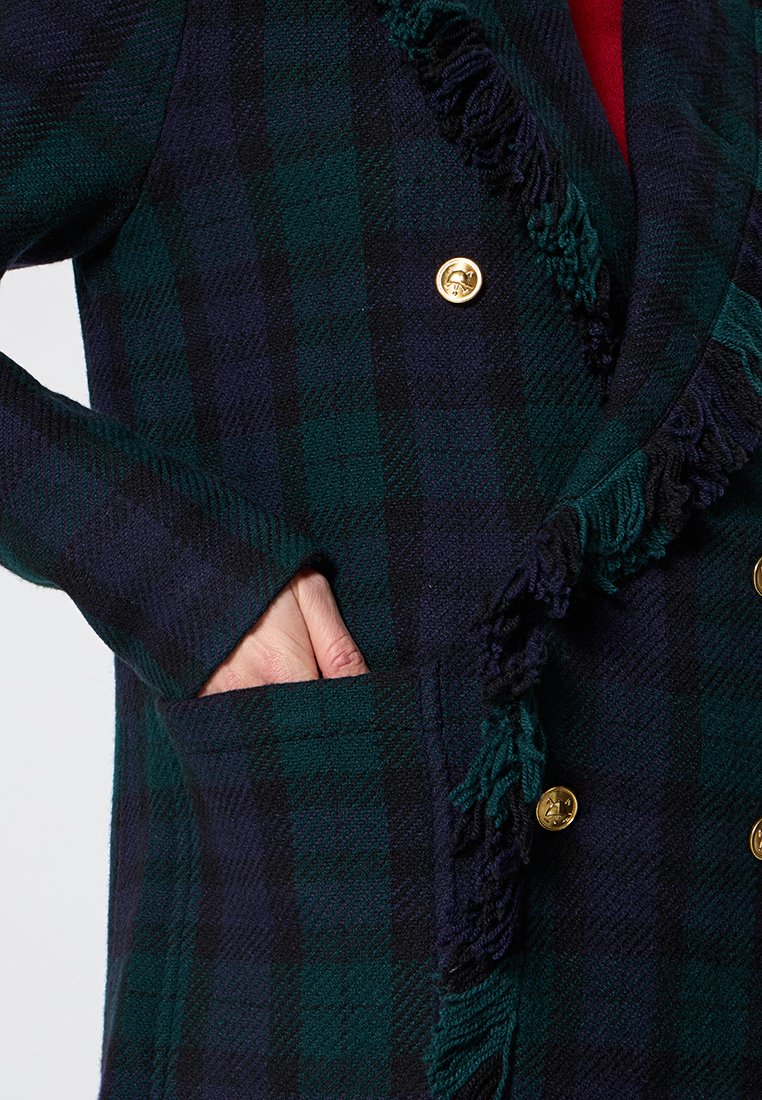 Podwójnie zapięty płaszcz w kratę w ciemnozielonym i granatowym kolorze z złotymi guzikami, teksturowana tkanina i frędzle wzdłuż kołnierza i kieszeni.