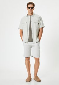 Chemise beige clair à manches courtes avec deux poches poitrine, t-shirt gris à col rond en dessous, short clair et chaussures à enfiler marron.