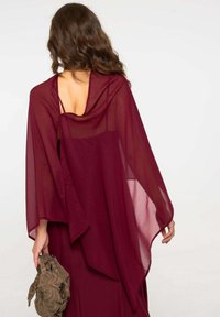Capa sheer color burdeos con espalda drapeada, diseño fluido y textura suave. Modelo sostiene un accesorio estampado en la mano.