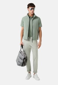 Lys grøn ærmeløs jakke over en matchende polo, lysegrå bukser og hvide sneakers. Holder en grå puffy taske med sorte stropper.
