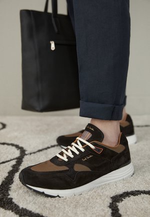 Schwarze und braune Sneakers mit weißen Schnürsenkeln, getragen zu hochgekrempelten dunkelblauen Hosen, stehend auf einem hellen Teppich neben einer schwarzen Tote-Tasche.