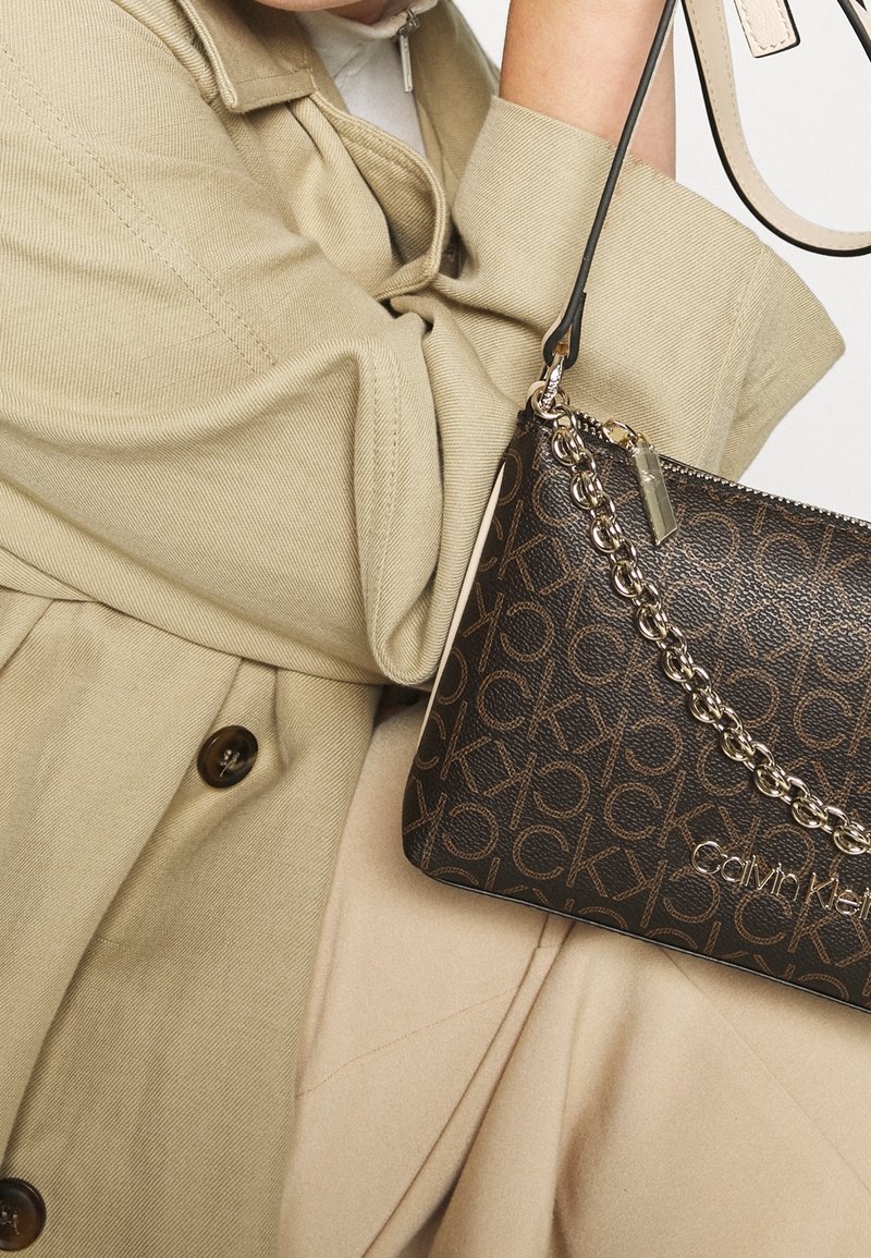 Femme tenant un sac bandoulière marron avec logo Calvin Klein et détail chaîne dorée, portant un trench beige et un pantalon clair.