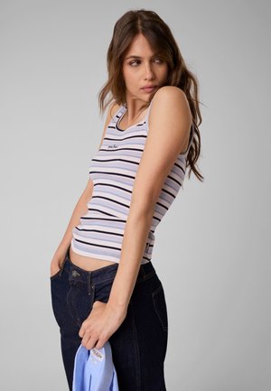 Jeune femme aux cheveux bruns portant un débardeur rayé blanc et bleu et un jean foncé, tenant un vêtement bleu clair dans une main.