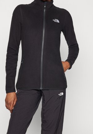 Personne portant une veste en polaire noire zippée et un pantalon assorti avec le logo The North Face, debout avec une main dans la poche.