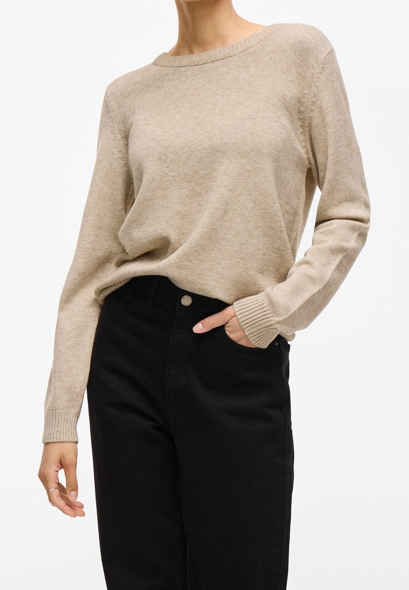 Femme portant un pull en maille beige à manches longues, rentré dans un pantalon noir taille haute, une main dans la poche et l'autre détendue le long du corps.