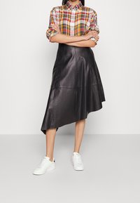 Person wearing a multicolored plaid shirt with rolled sleeves, black asymmetrical leather skirt, and white sneakers standing with arms crossed.