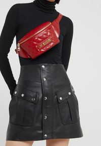 Červená prošívaná ledvinka se zlatým logem, má zipové uzavření. Černá kožená mini sukně s předními patentkami a kapsami, texturovaný pas.