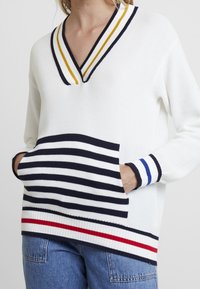 Vit stickad tröja med v-ringning, randiga detaljer i marinblått, gult och rött, med en framficka med horisontella ränder och ribbade ärmslut.