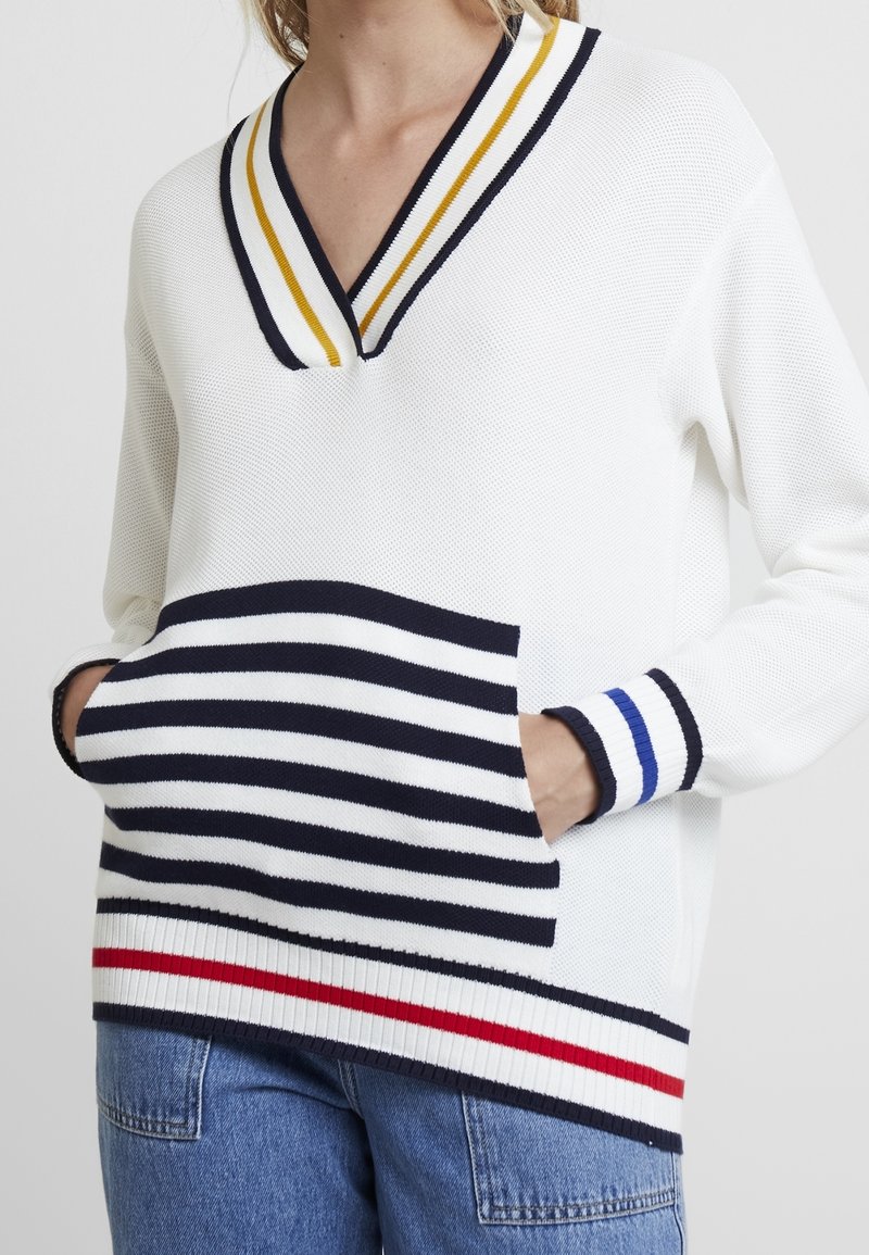 Vit stickad tröja med v-ringning, randiga detaljer i marinblått, gult och rött, med en framficka med horisontella ränder och ribbade ärmslut.