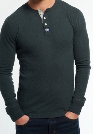 Hombre vestido con una camiseta henley de manga larga verde oscuro, con una tapeta de botones gris y un pequeño parche con logo en el pecho, con las manos en los bolsillos.