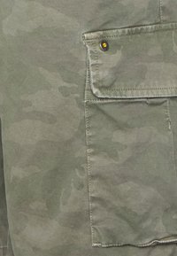 Tkanina s kamuflážním vzorem v odstínech zelené, se záplatovou kapsou a černým knoflíkem. Hladká textura s viditelnými detaily prošívání.