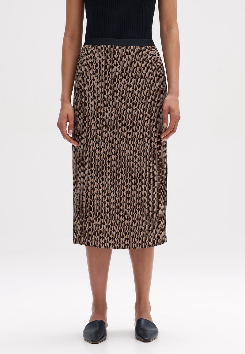 Femme portant une jupe midi à imprimé géométrique marron et noir, associée à un top noir sans manches et des ballerines noires pointues.