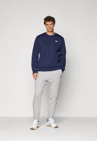 Granatowy sweatshirt z okrągłym dekoltem i ściągaczami u rękawów, w zestawie z jasnoszarymi spodniami dresowymi. Oba elementy mają logo na klatce piersiowej i nogawkach. Białe sneakersy.