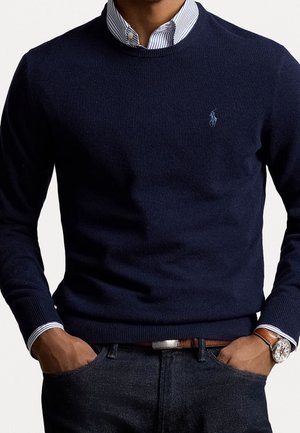 Mężczyzna w granatowym swetrze nałożonym na koszulę w paski z kołnierzykiem, z rękami w kieszeniach i zegarkiem na lewej ręce.