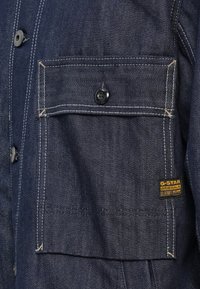Donkerblauwe denimjaszak met zwarte knop en gele G-STAR Originals RAW-label gestikt aan de zijkant.