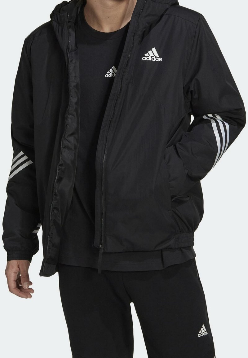Chaqueta negra ligera con capucha, cremalleras y acentos de tres rayas blancas en las mangas. Combinada con una camiseta y pantalones negros.