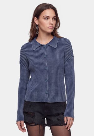 Donna con un maglione blu a coste con bottoni e colletto e pantaloncini di jeans neri strappati su uno sfondo semplice.