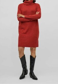 Robe-pull rouge à col roulé en tissu maille doux, dotée de manches longues et d'une coupe ample. Associée à des bottes en cuir noires montant jusqu'aux genoux.