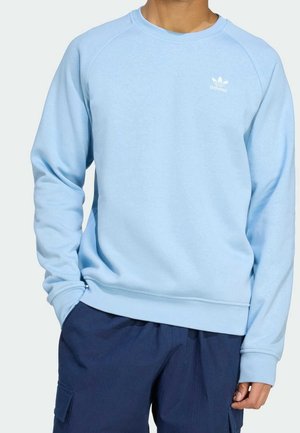 Mężczyzna w jasnoniebieskiej bluzie Adidas z logo na piersi oraz ciemnoniebieskich spodniach bojówkach, stojący z jedną ręką w kieszeni.