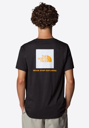 Mand med krøllet hår iført sort The North Face t-shirt med orange logo og tekst "Never Stop Exploring" på ryggen, stående mod hvid baggrund.
