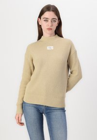 Beżowy sweter z prążkowanego dzianiny z wysokim kołnierzem, długimi rękawami i kwadratową etykietą materiałową na piersi. W zestawie z niebieskimi dżinsami.