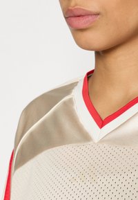 Camiseta deportiva color tostado con detalles en rojo, que presenta un escote en V, textura de malla transpirable y un patrón de pequeñas perforaciones por todo el tejido.