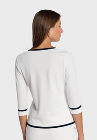 Haut blanc à manches trois-quarts, bordure marine au niveau du col et des poignets, texture lisse, coupe ajustée. Aucun détail supplémentaire présent.