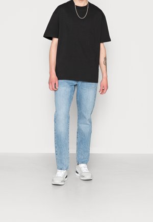 T-shirt nera in cotone con maniche corte, abbinata a jeans in denim azzurro chiaro. Le sneakers sono grigie con dettagli bianchi. Design semplice e casual.