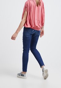 Luźna różowa bluzka z marszczonymi ramionami, zestawiona z ciemnoniebieskimi skinny jeansami i szarymi wzorzystymi sneakersami. Widok z tyłu na białym tle.