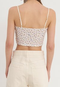 Top court floral blanc avec des fines bretelles, en matière texturée et muni d'une coupe ajustée ; associé à un jean beige taille haute avec des poches arrière.