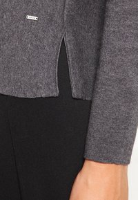 Pull en maille grise avec fentes latérales et une étiquette de marque discrète. Texture douce et manches ajustées, associé à un bas noir.