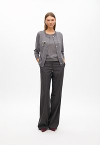 Szary kardigan z okrągłym dekoltem i dekoracyjną broszką; nałożony na szary top; zestawiony z szerokimi spodniami w prążki i fioletowymi butami.