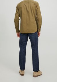 Olivgrünes Hemd mit langen Ärmeln, Baumwollmaterial, Rückenpasse, abgerundetem Saum, kombiniert mit blauen Jeans und hellen Schuhen.