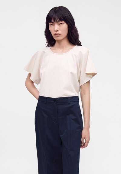 Femme aux longs cheveux noirs portant un haut beige à manches courtes et larges, et un pantalon bleu marine taille haute, debout devant un fond blanc.