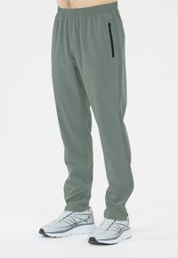 Rekbare, lichtgewicht grijs-groene sportbroek met een elastische tailleband en een ritssluitingzak aan de zijkant, gecombineerd met grijze sneakers.