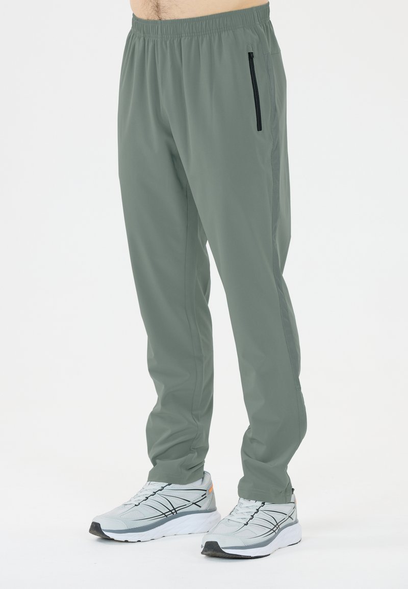 Rekbare, lichtgewicht grijs-groene sportbroek met een elastische tailleband en een ritssluitingzak aan de zijkant, gecombineerd met grijze sneakers.