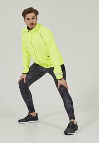 Veste de sport jaune fluo avec accents réfléchissants, leggins noirs à motifs et chaussures de sport noires en position d'étirement sur un fond uni.