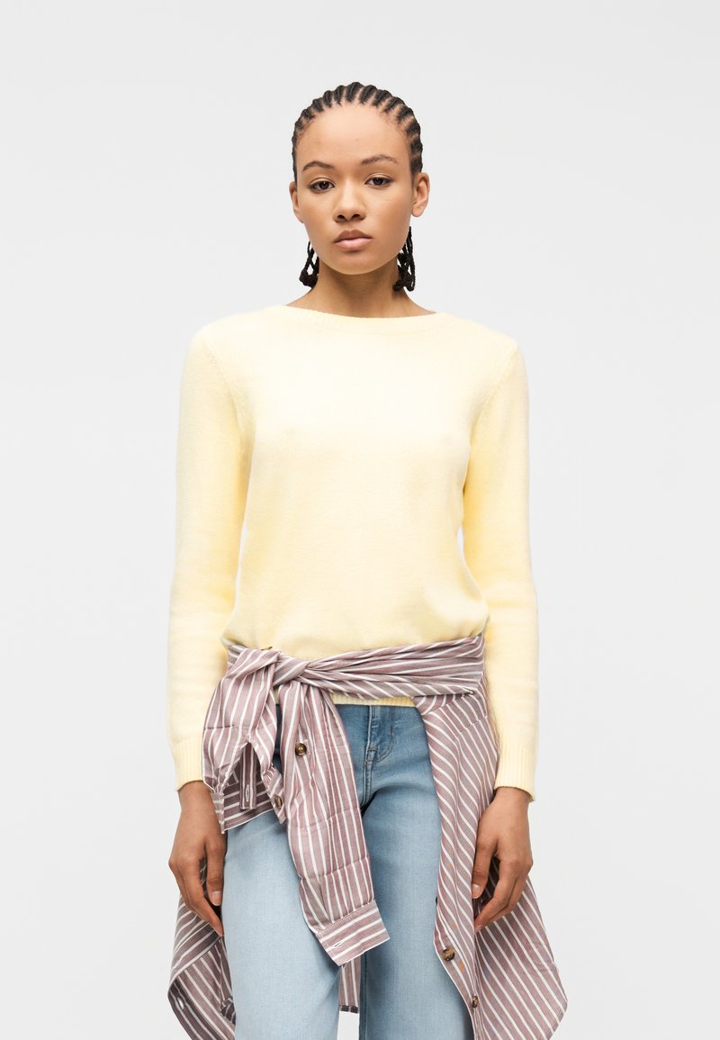 Jeune femme aux cheveux tressés portant un pull jaune pâle, un jean bleu clair et une chemise rayée nouée autour de la taille, sur un fond uni.